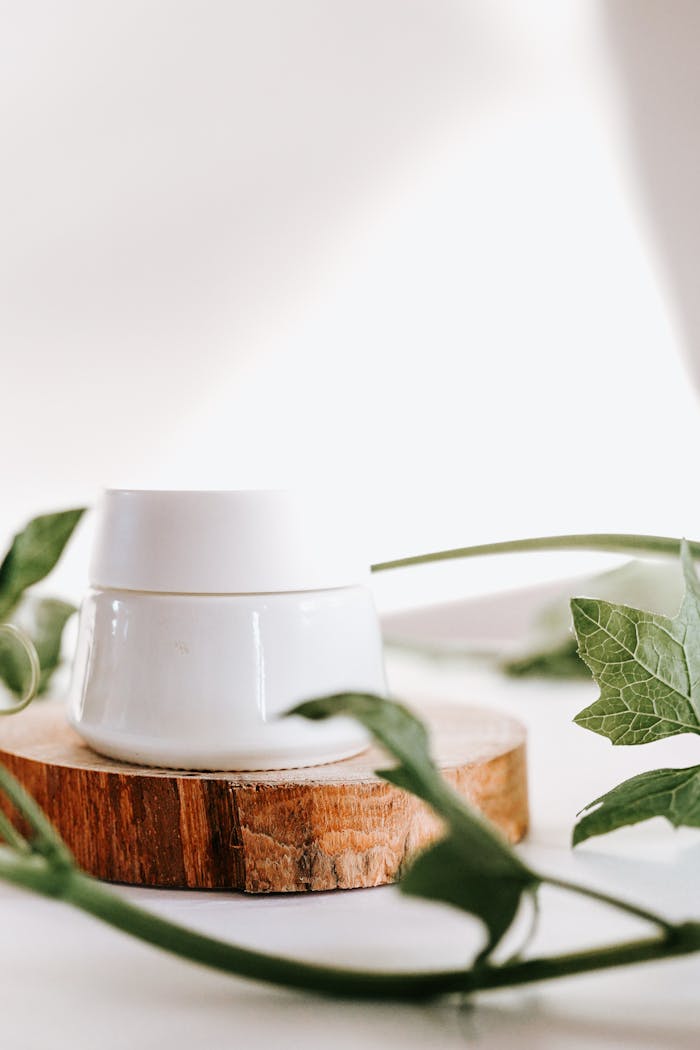 Elegant skincare jar placed on wooden slab surrounded by green leaves.
